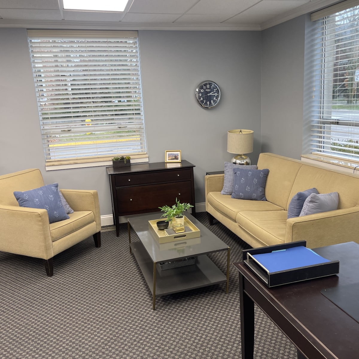 A modern office waiting area with a beige couch, a matching armchair, a coffee table, and a dark wooden cabinet, all arranged near large windows with blinds.