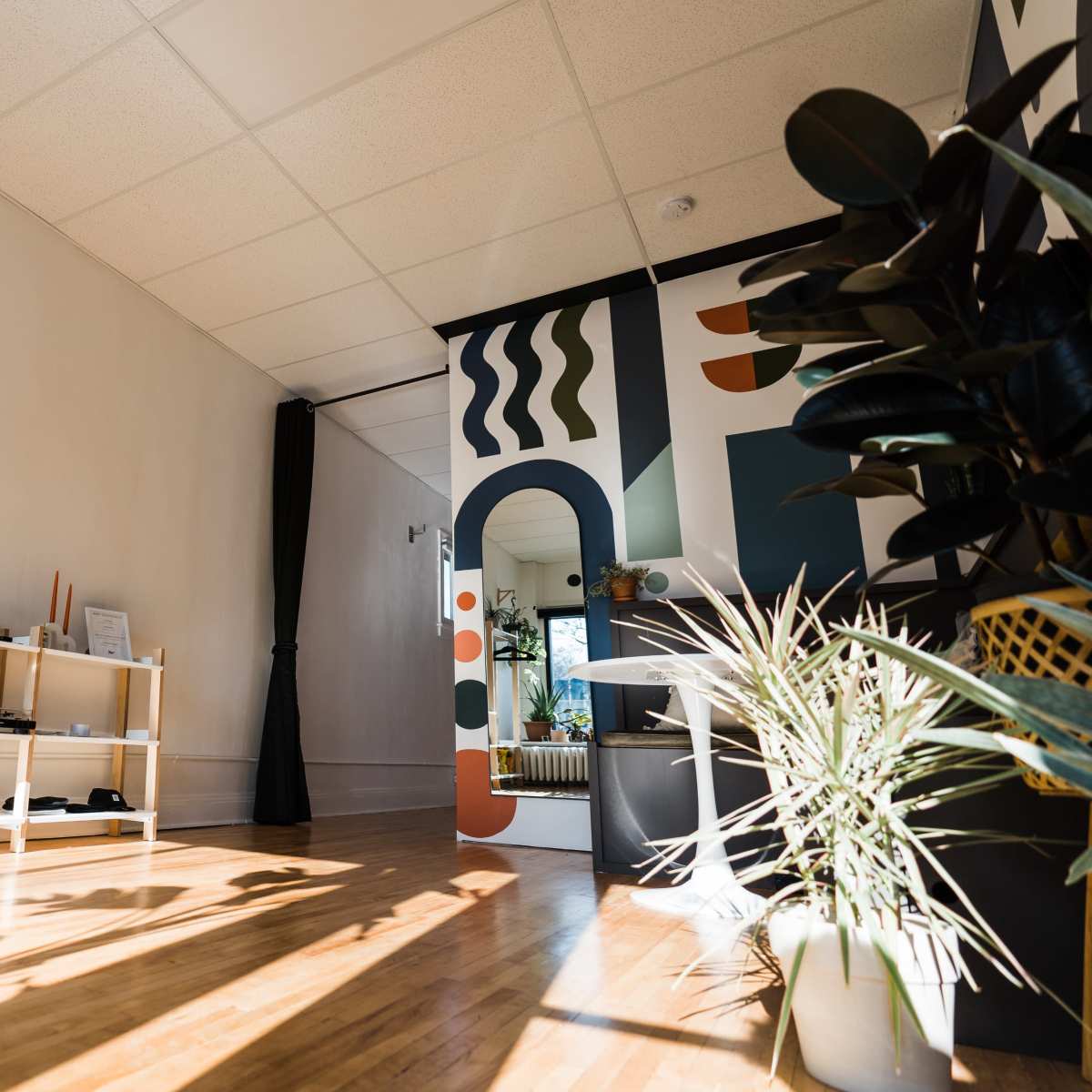 A well-lit room features wooden floors, shelves with books and decor, and a wall with geometric artwork.