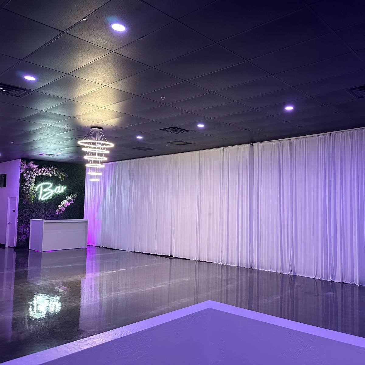 An empty indoor event space featuring a bar area, white drapery along one wall, and purple lighting.
