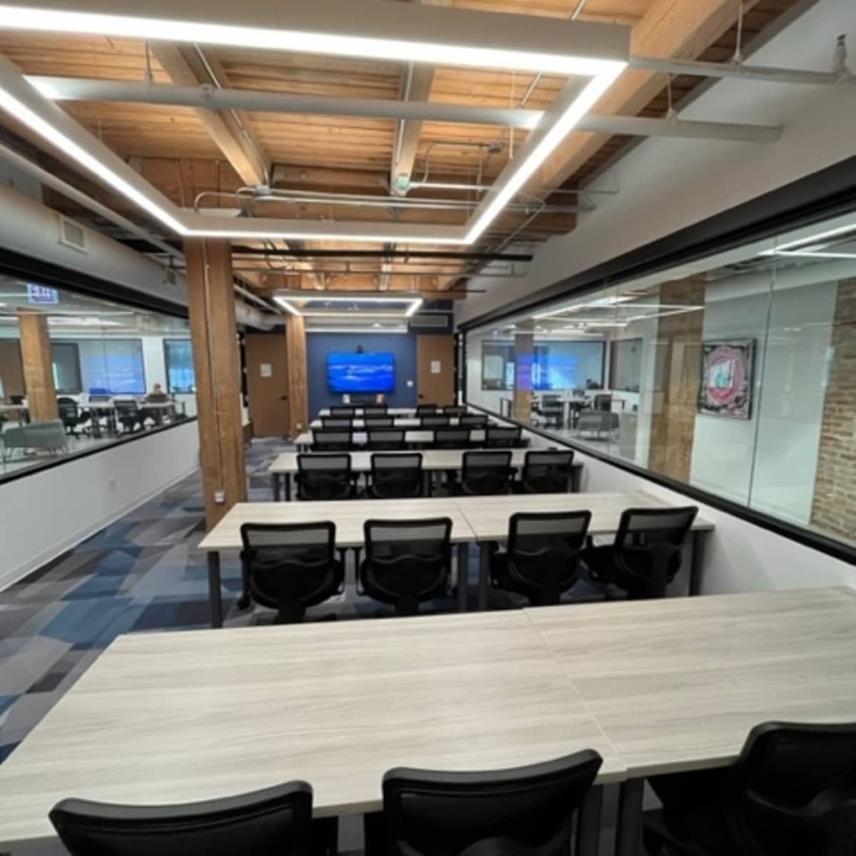 A modern office space featuring rows of tables and ergonomic chairs, with large windows and exposed wooden beams overhead.