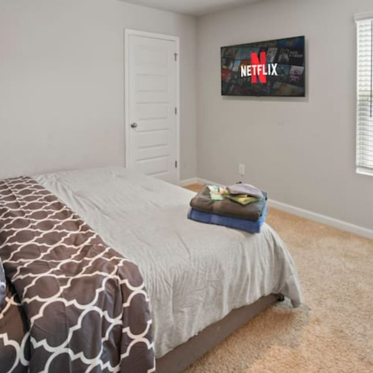 A neatly made bed with patterned bedding is positioned in a room featuring a television displaying the Netflix logo and a window with blinds.