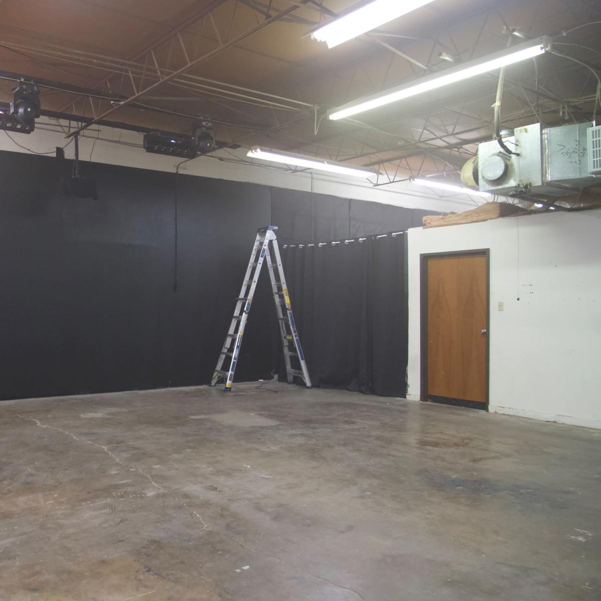 An empty room with a black wall, a ladder, and a door, illuminated by overhead lights.