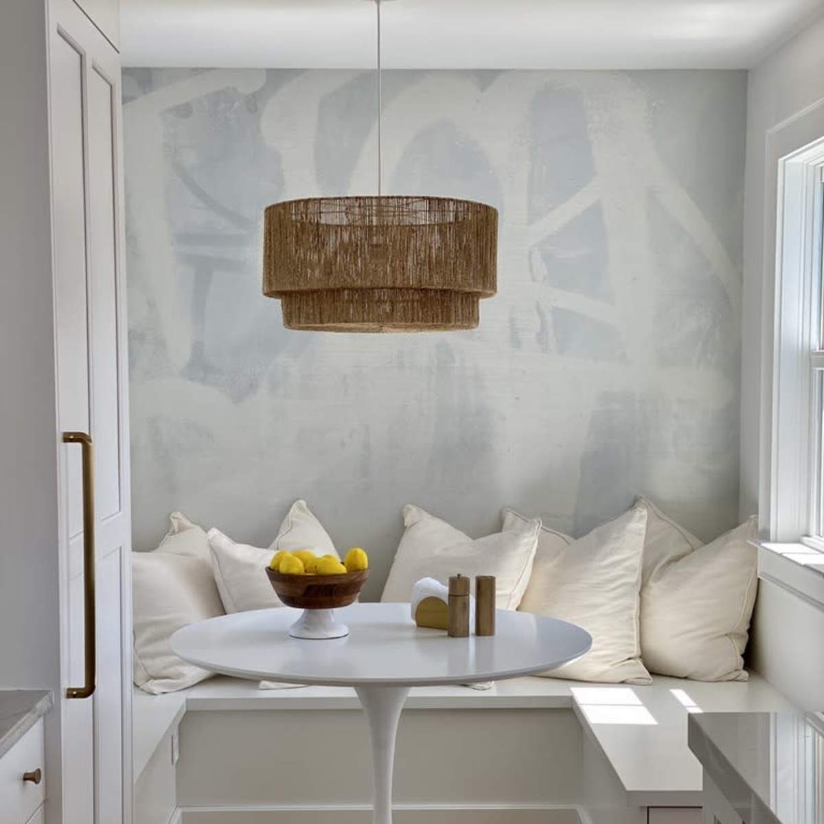 A cozy breakfast nook featuring a round white table, a bowl of lemons, and a built-in bench with white pillows against a softly painted wall.