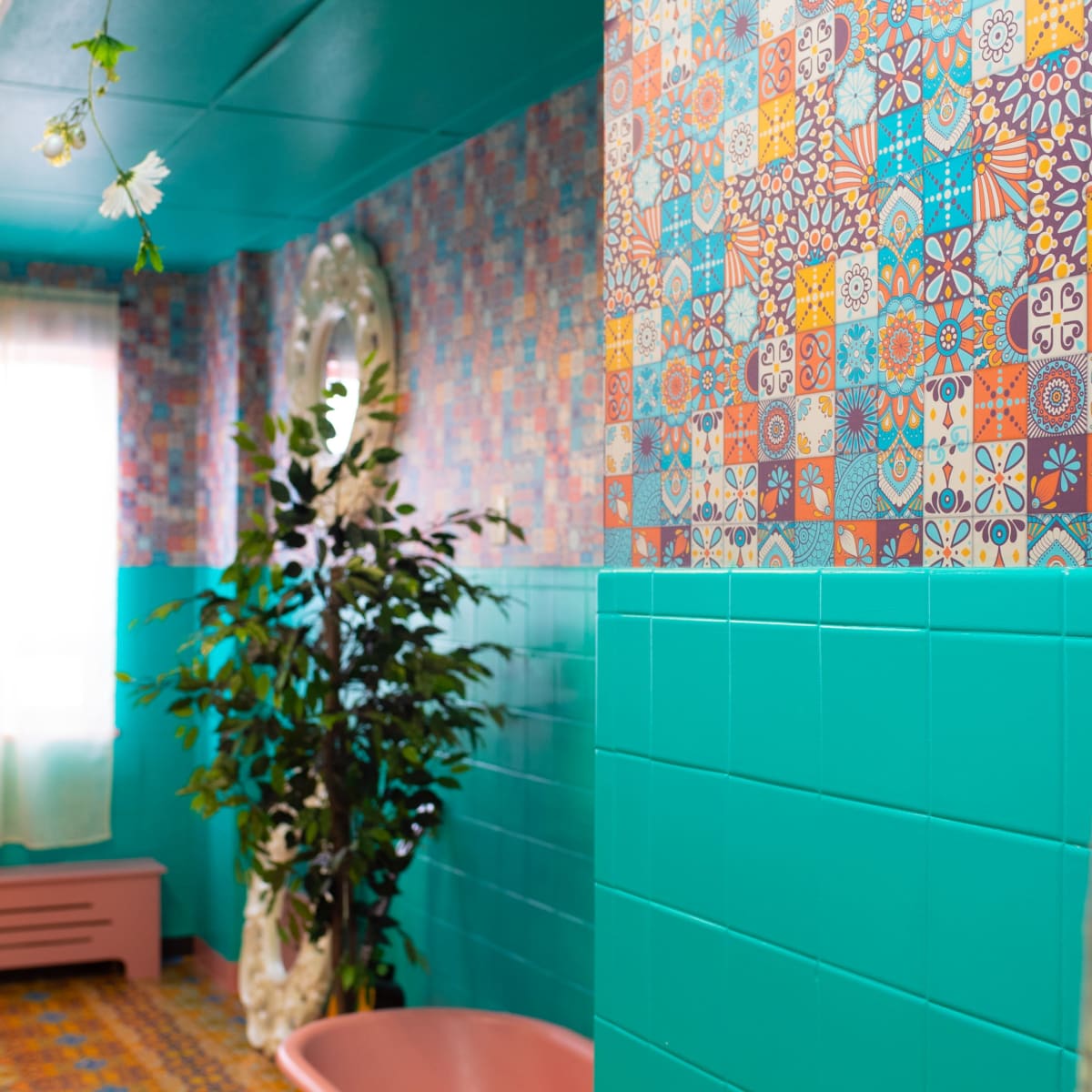 A brightly colored bathroom features patterned tile walls, a pink bathtub, and a plant in the corner, with sunlight streaming through sheer curtains.