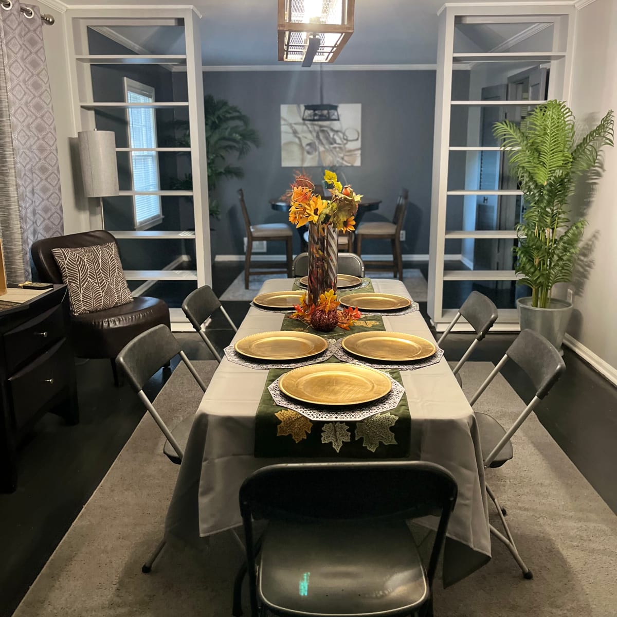 A dining room is set up with a large table adorned with plates and a centerpiece of autumn flowers.