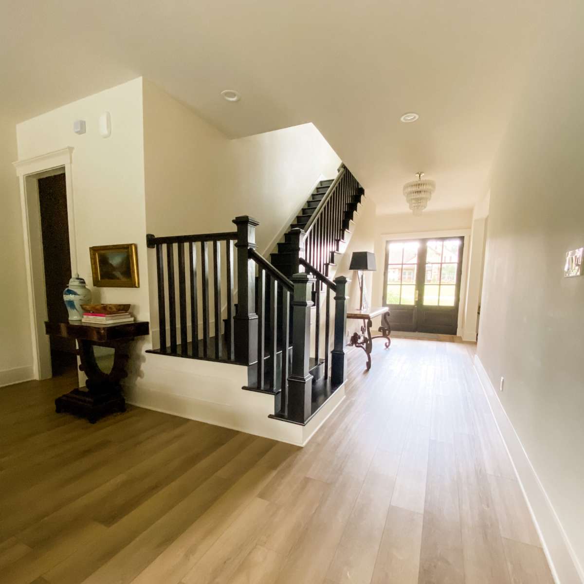 A bright indoor hallway featuring a staircase with black railings, wooden flooring, and a foyer area with a side table and decorative items.