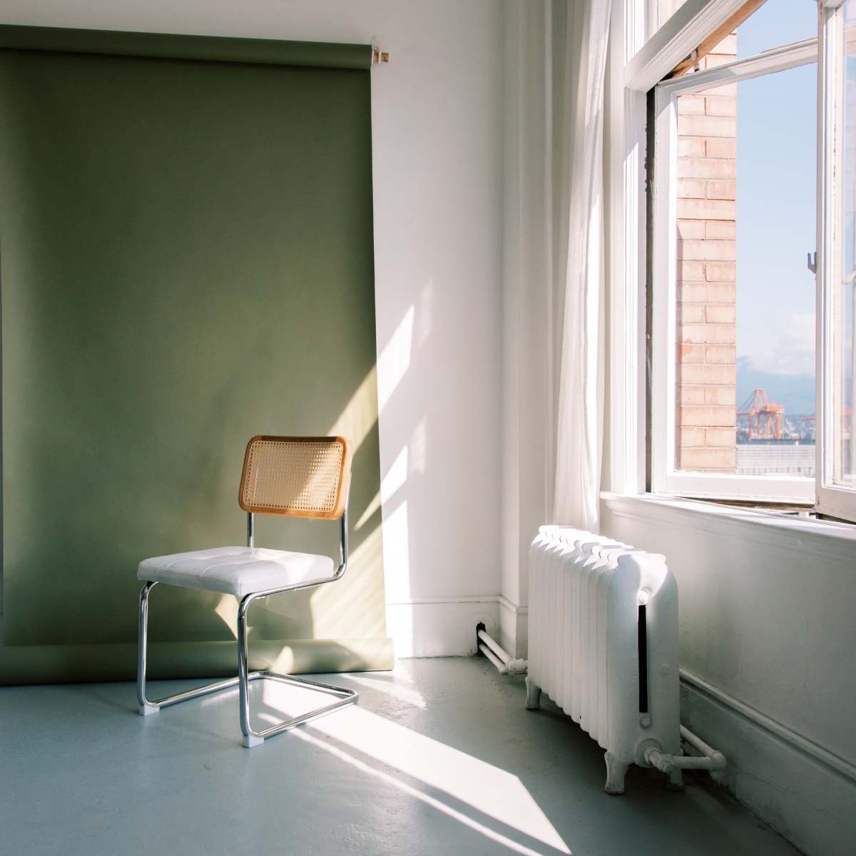 A minimalist room features a single chair in front of a green backdrop, with sunlight streaming in through a window.