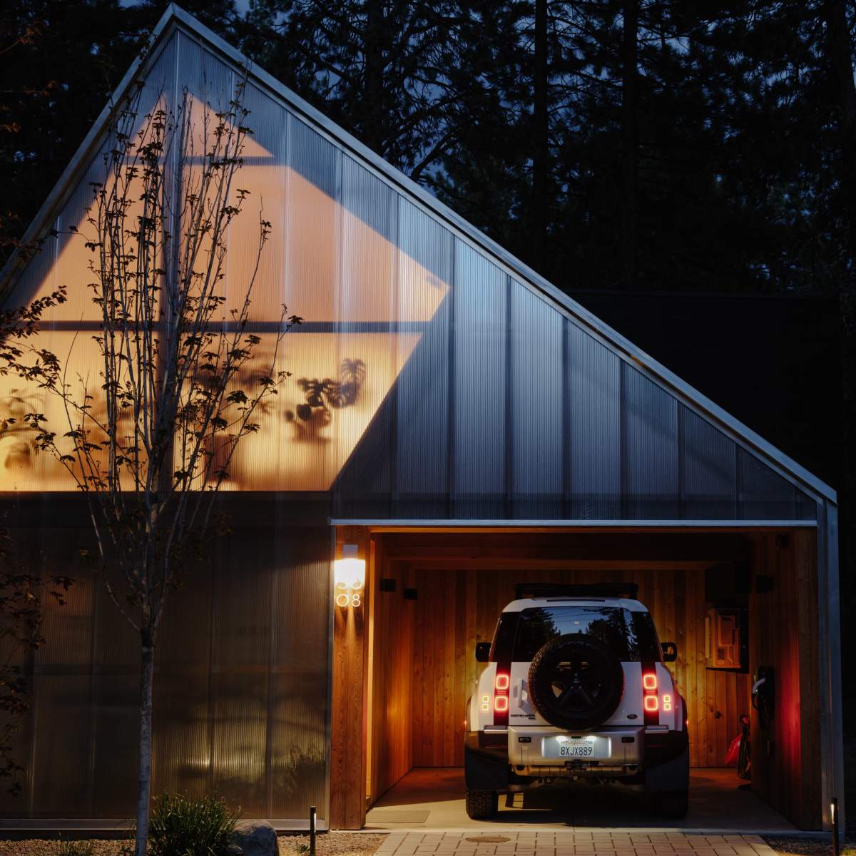 A vehicle is parked inside a carport of a modern house illuminated by warm lights at dusk.