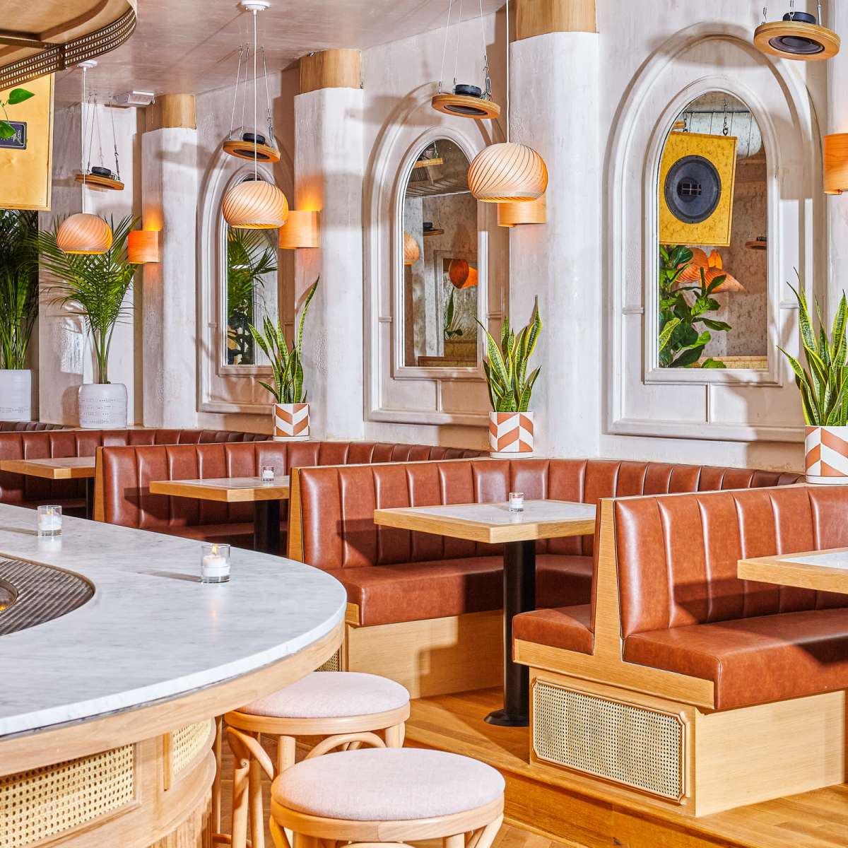 A modern dining space with leather banquette seating, white walls, and plants, illuminated by pendant lights above small tables.
