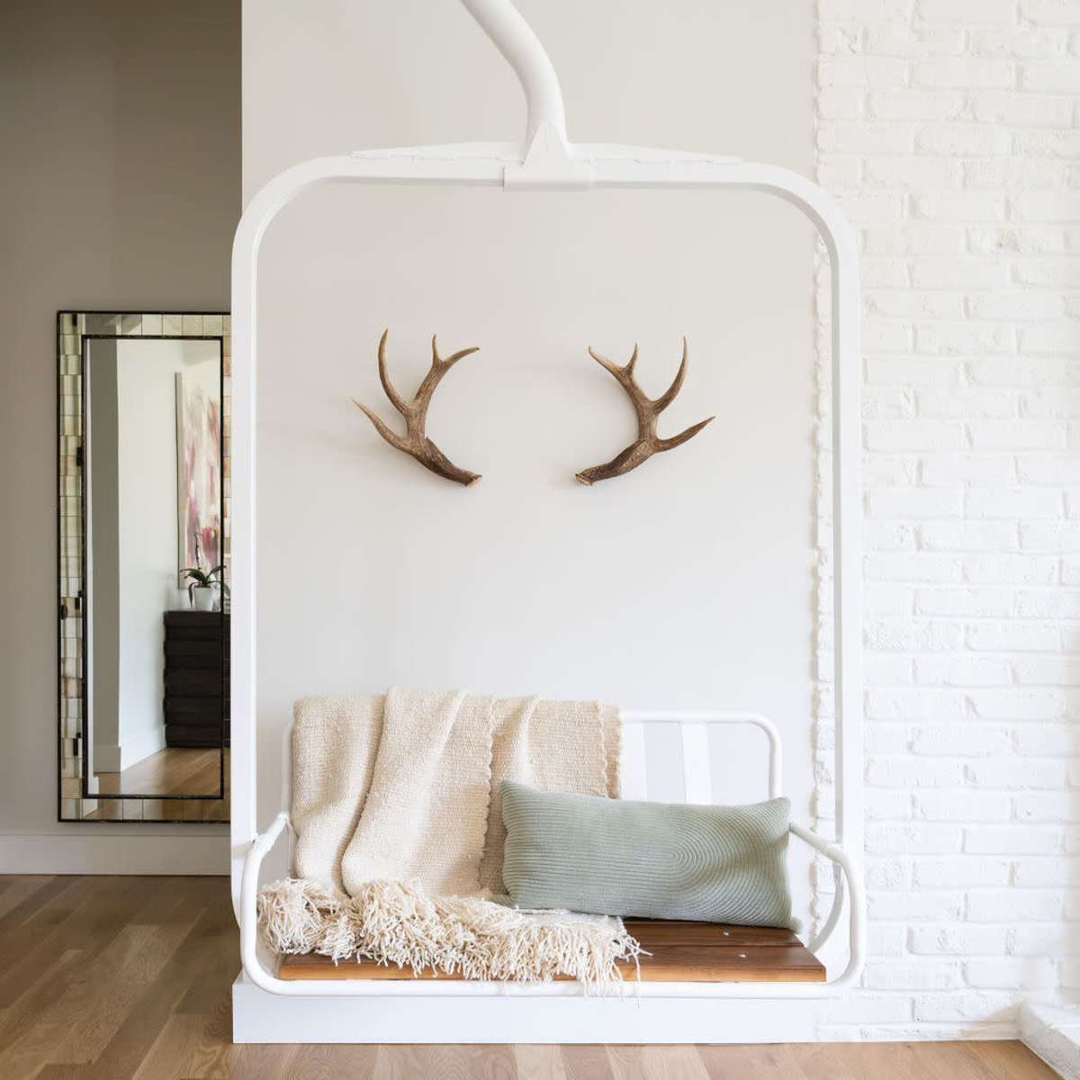 A white swing is mounted against a textured wall, adorned with deer antlers, a neutral blanket, and a green cushion, with a mirror reflecting the space.