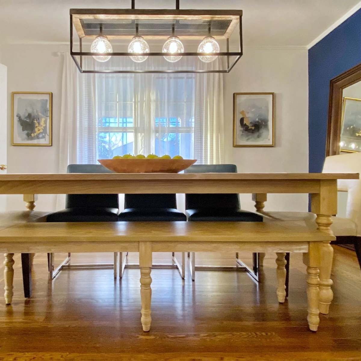 A wooden dining table with a bowl of fruit is set beneath a modern light fixture in a room featuring blue and white walls.