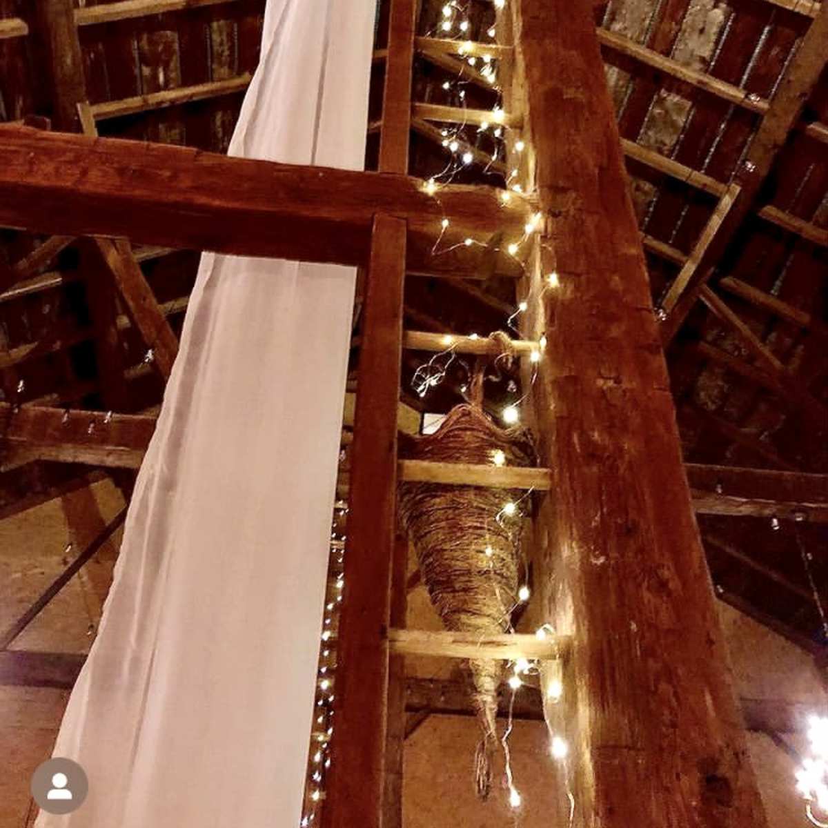A wooden ladder is positioned against a rustic beam adorned with soft drapery and string lights in a barn setting.