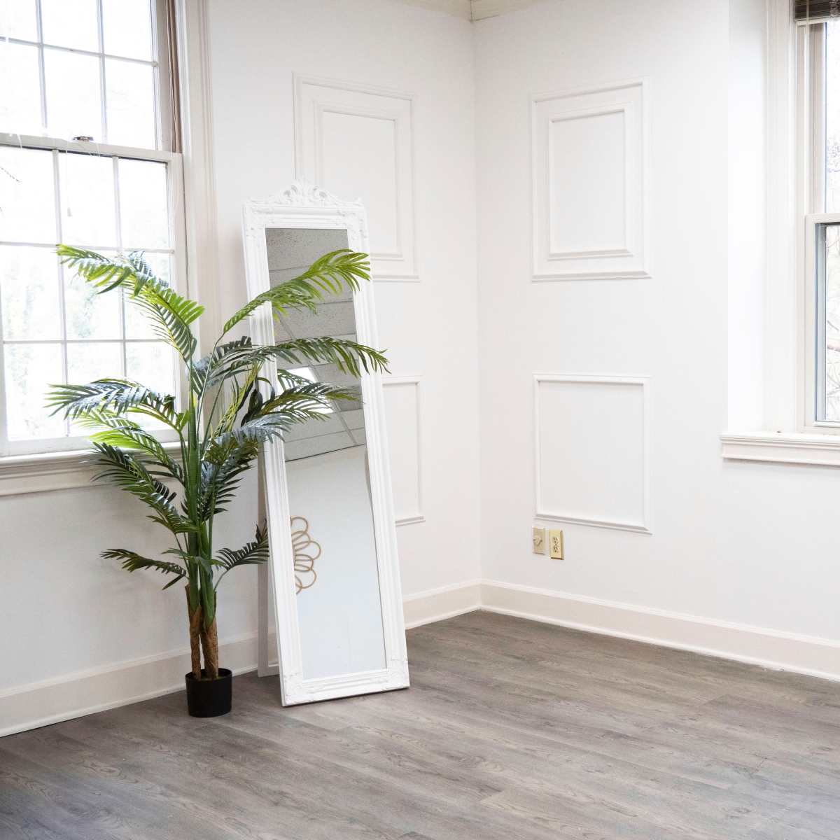 A minimalist room with a large mirror leaning against the wall next to a potted plant, featuring white walls and windows letting in natural light.
