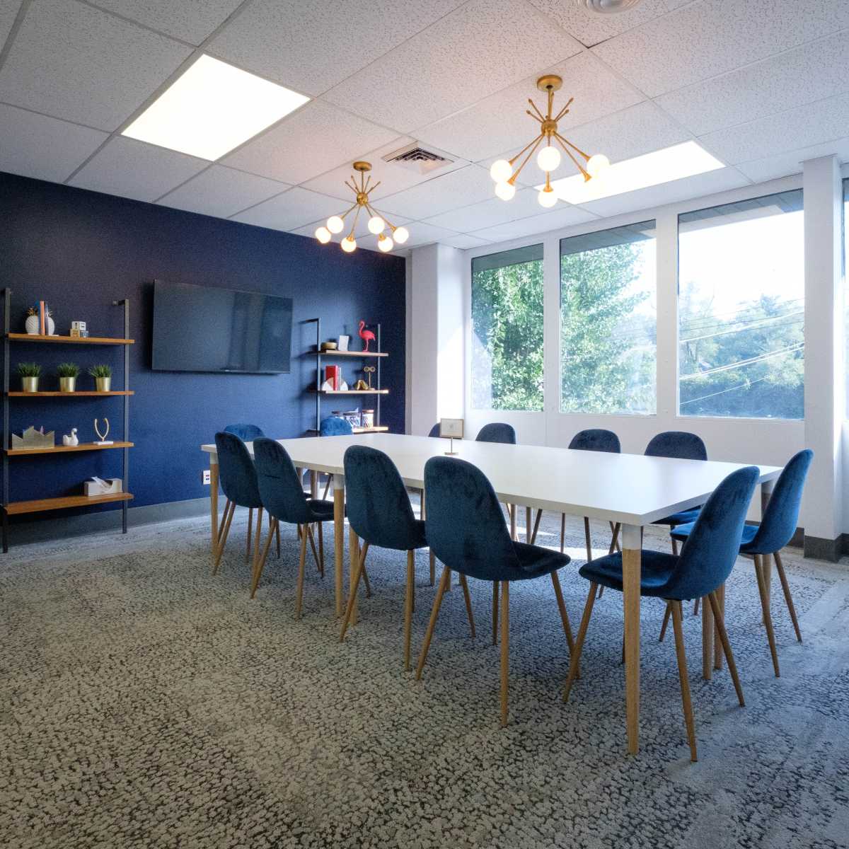 A modern conference room featuring a long white table surrounded by blue chairs, with large windows allowing natural light and a television mounted on the wall.