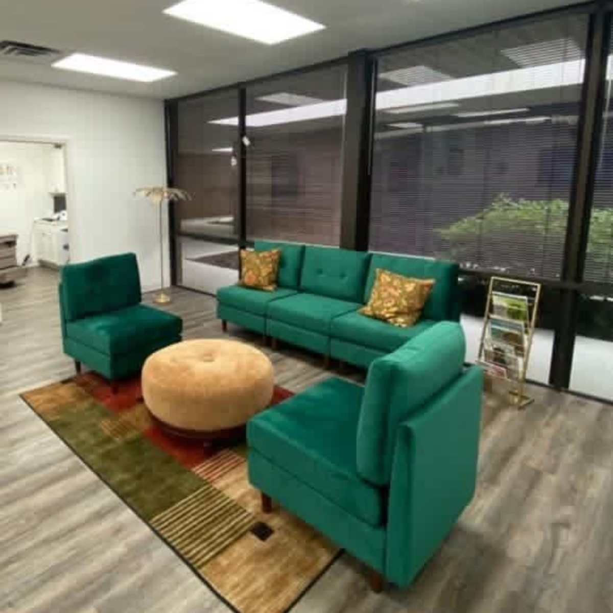 A modern sitting area featuring a green sofa with decorative pillows, two matching chairs, a round ottoman, and large windows letting in natural light.