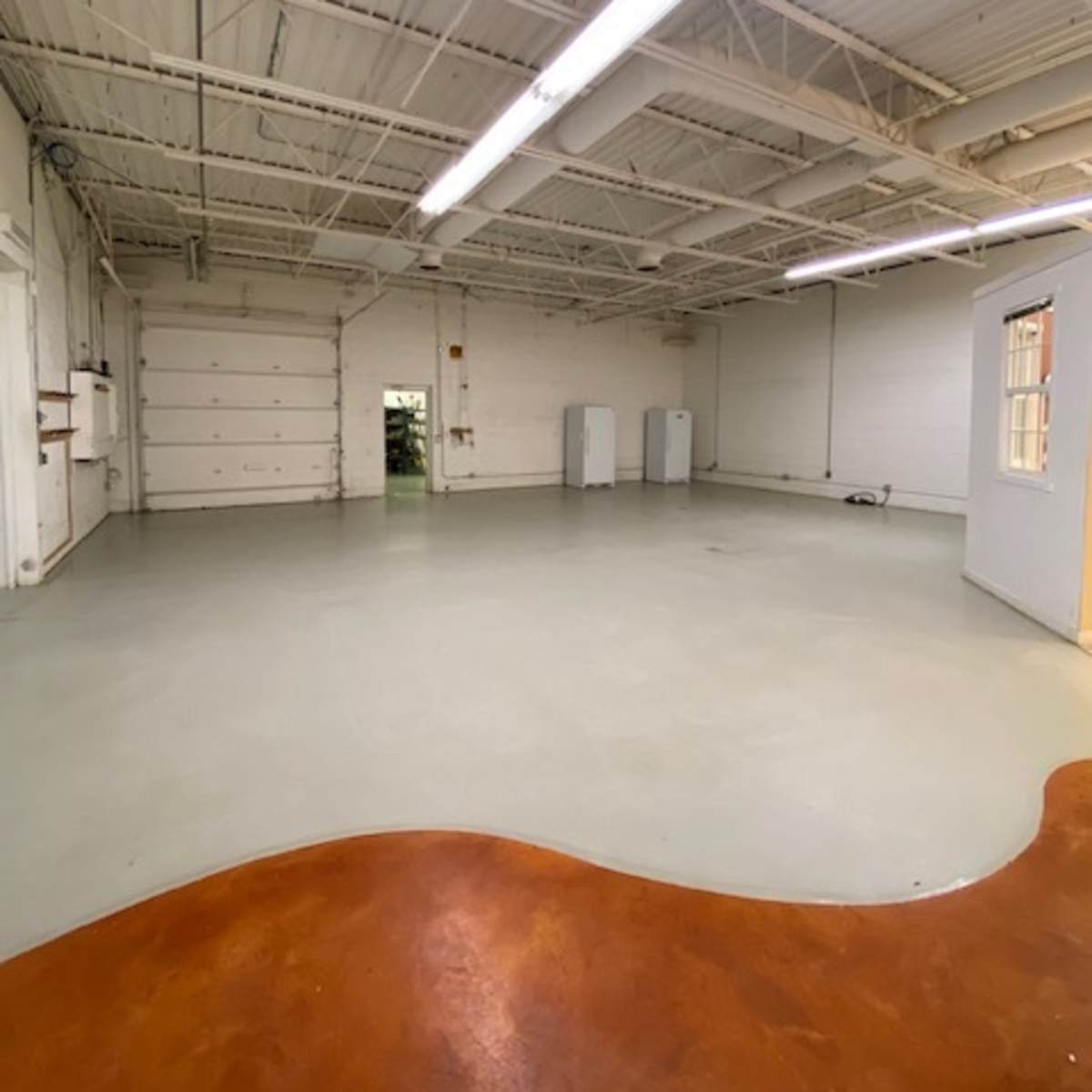 A spacious, empty room with a smooth floor and white walls, featuring overhead lighting and a few appliances against one wall.