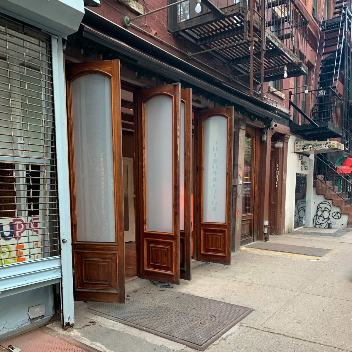 A series of wooden double doors standing open on a sidewalk next to a building with fire escapes.