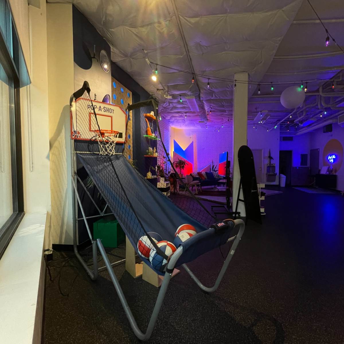 A recreational room featuring a basketball hoop, a lounge chair with basketballs, and colorful lighting in the background.
