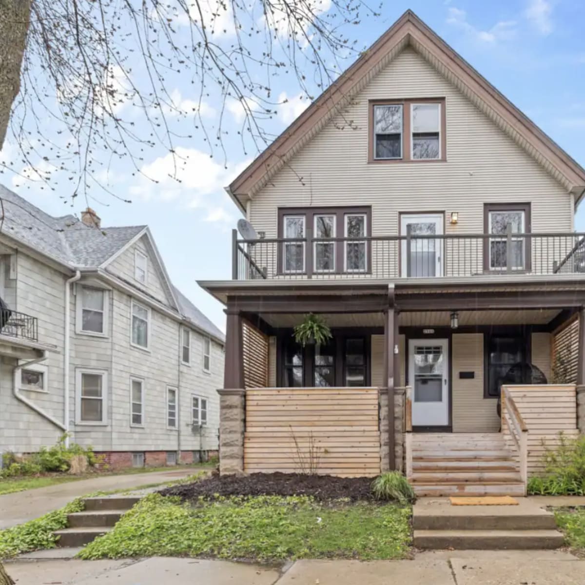 A three-story house with a front porch, wooden railings, and landscaped steps is situated on a tree-lined street.
