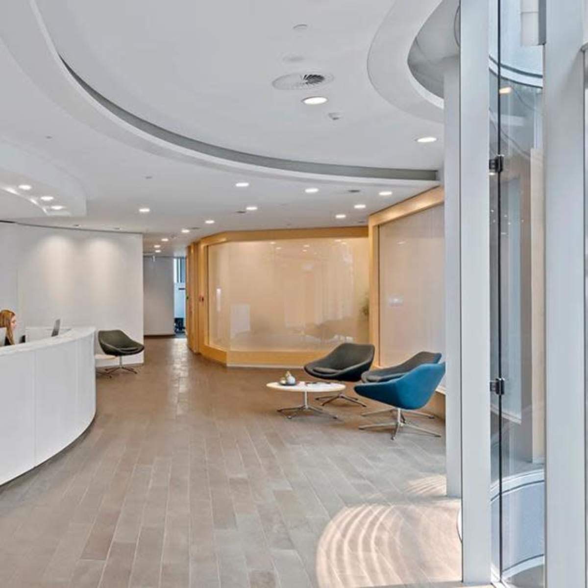 A modern reception area featuring a curved white front desk, two women seated at computers, and contemporary seating arrangements in a well-lit space.