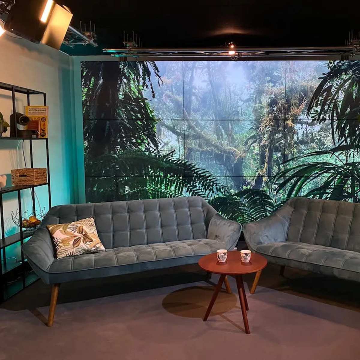 A modern studio setup with two teal sofas, a round wooden table in between, and a large backdrop featuring a tropical forest scene.