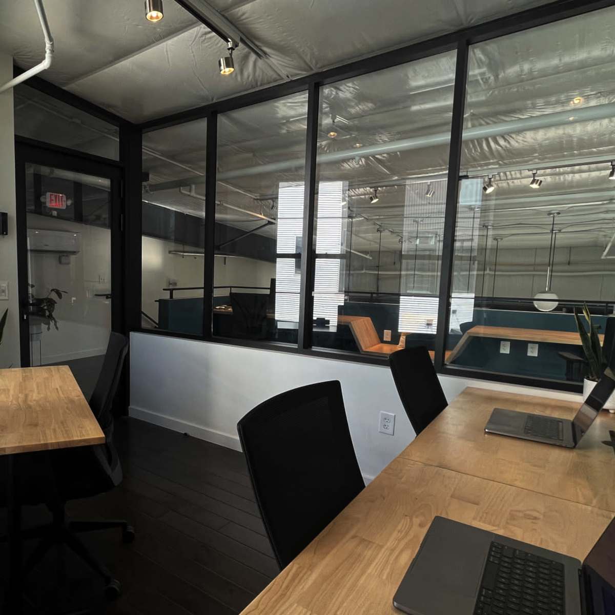 A modern office space with large windows, a wooden desk, and ergonomic chairs, featuring an open area visible through a glass partition.