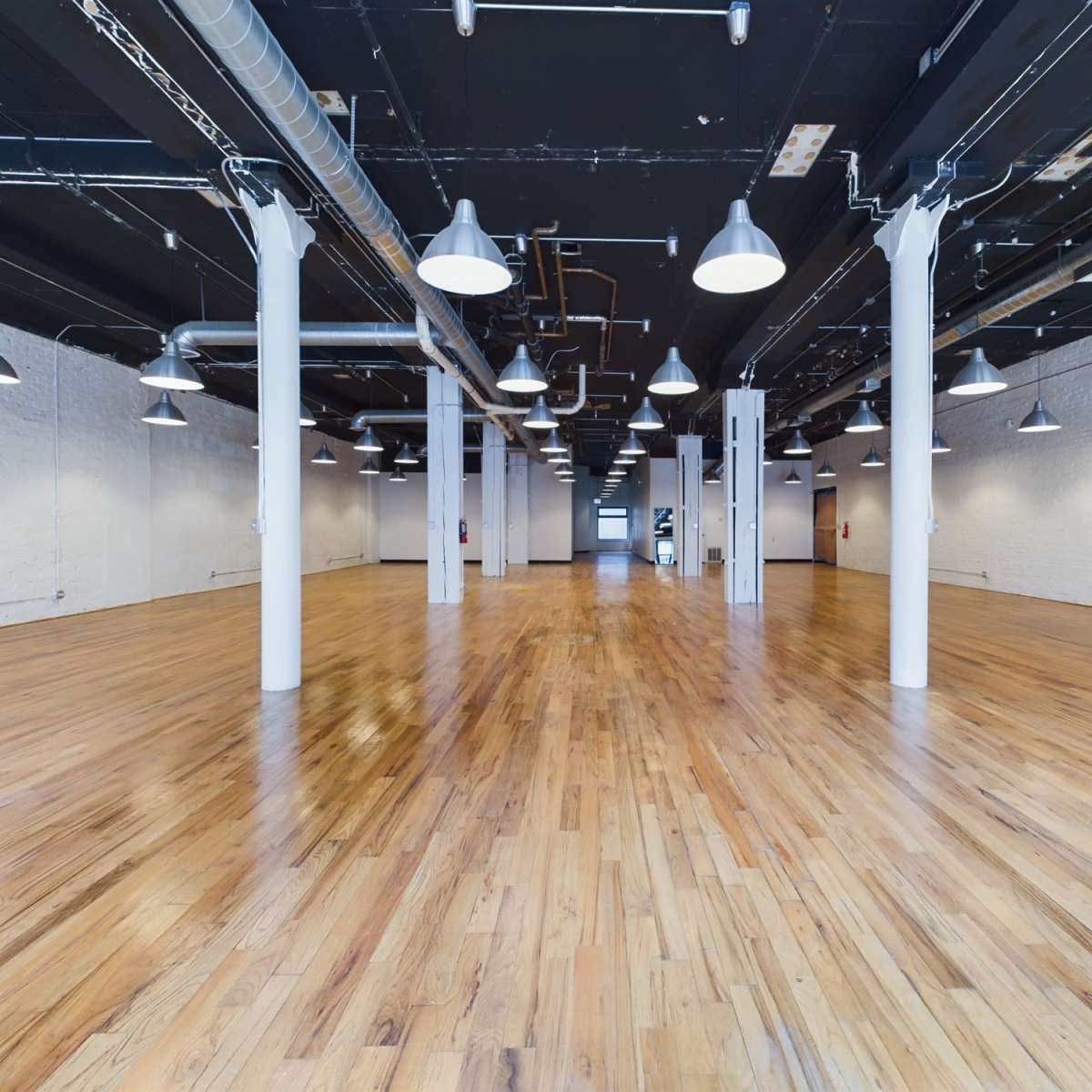 A spacious, empty room with polished wooden floors and exposed ductwork, illuminated by pendant lights.