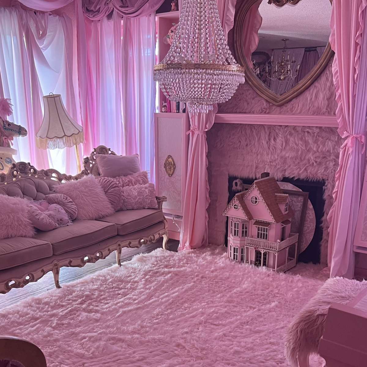 The room features a pink-themed decor with a chandelier, plush sofa, and a dollhouse near a plush rug.