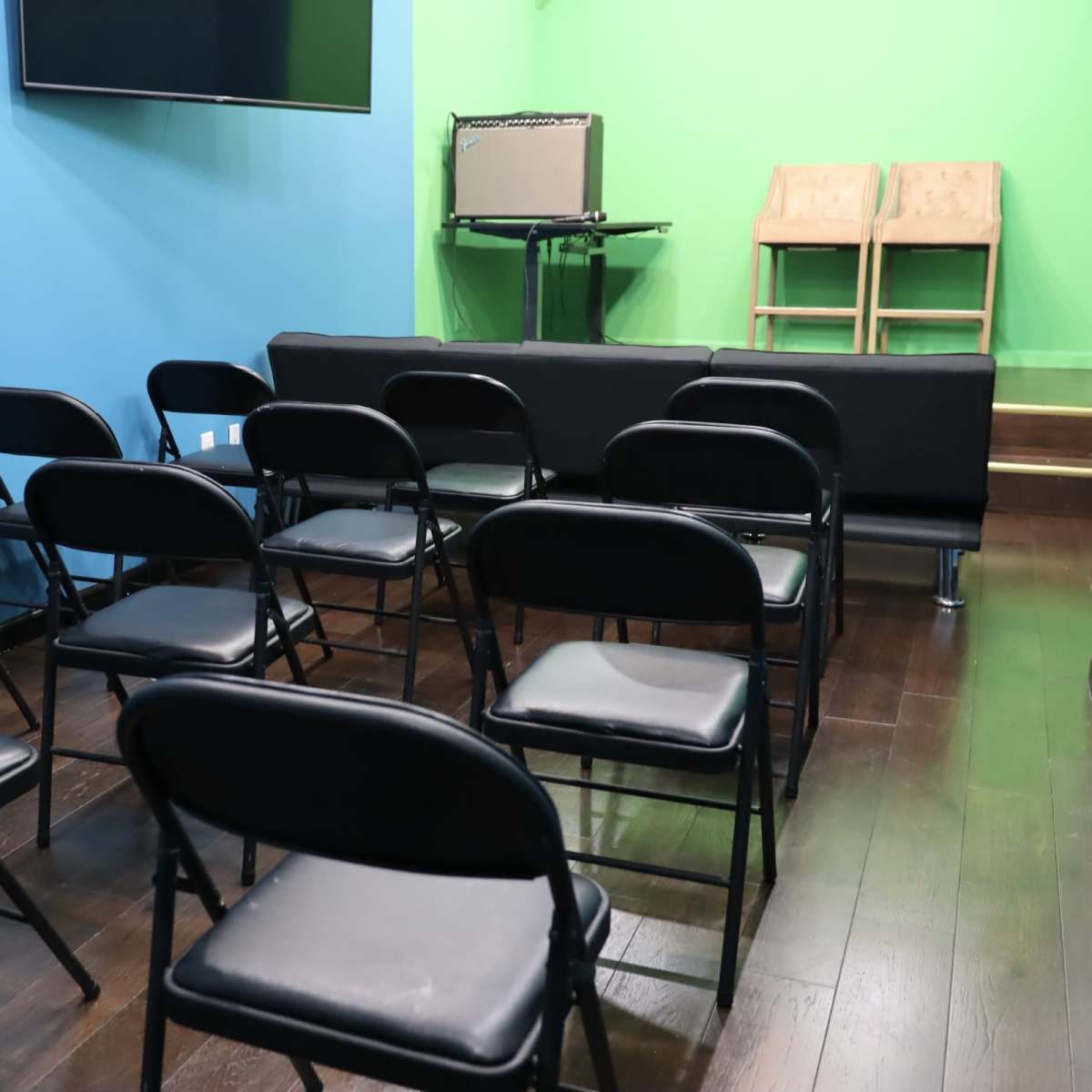 A small room with black folding chairs arranged in rows, facing a stage with an amplifier and two wooden chairs against a bright green backdrop.