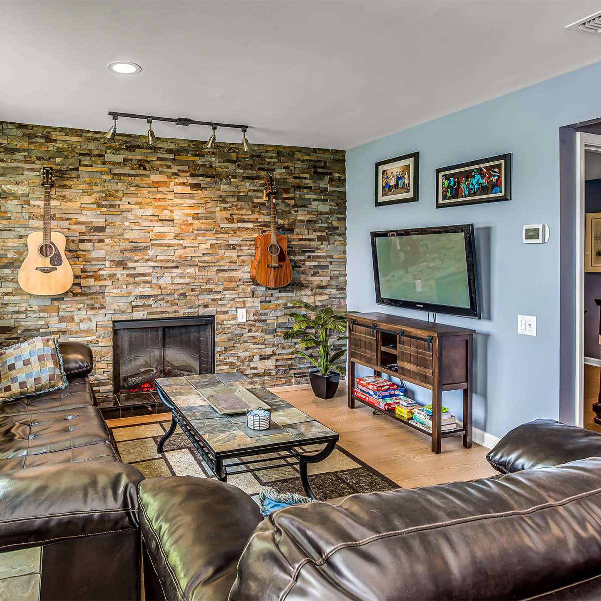 A living room with a stone accent wall featuring two guitars, a television on a stand, and a leather sectional sofa arranged around a coffee table.