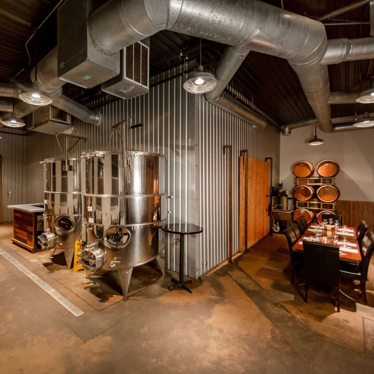 An industrial-style interior featuring metal fermentation tanks, wooden barrels, and a dining area with a table set for multiple guests.