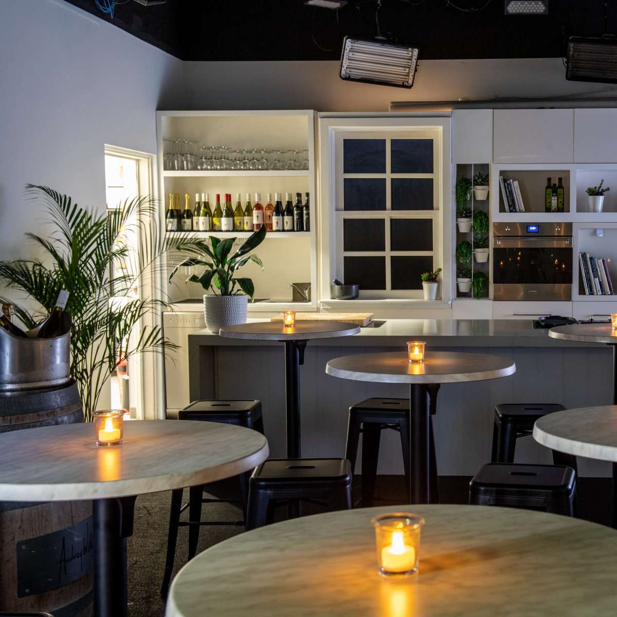 A modern restaurant interior with round tables, each featuring a lit candle, and a bar area with a backdrop of shelves displaying bottles and plants.