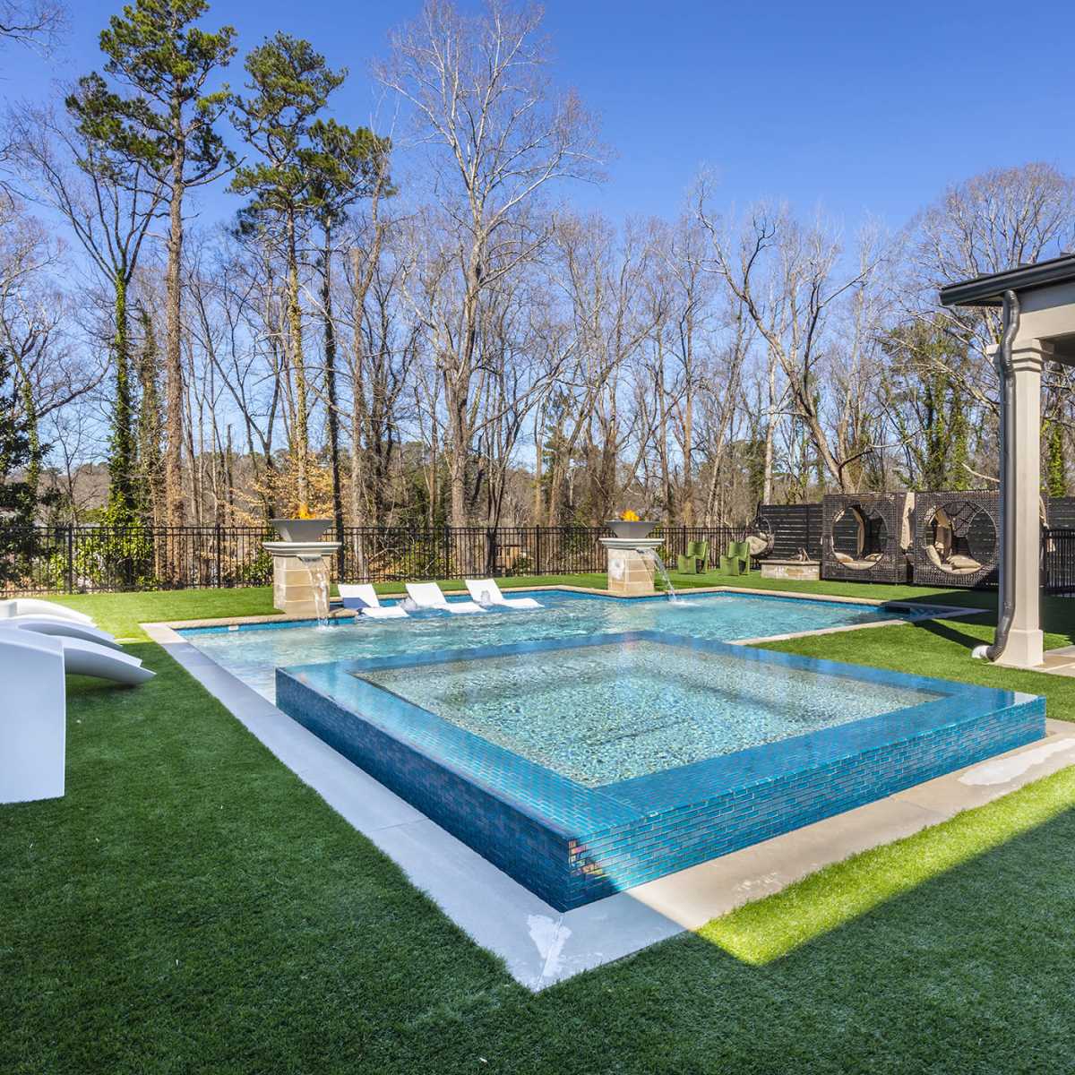 A rectangular swimming pool with a raised spa surrounded by green grass and lounge chairs in a wooded outdoor setting.