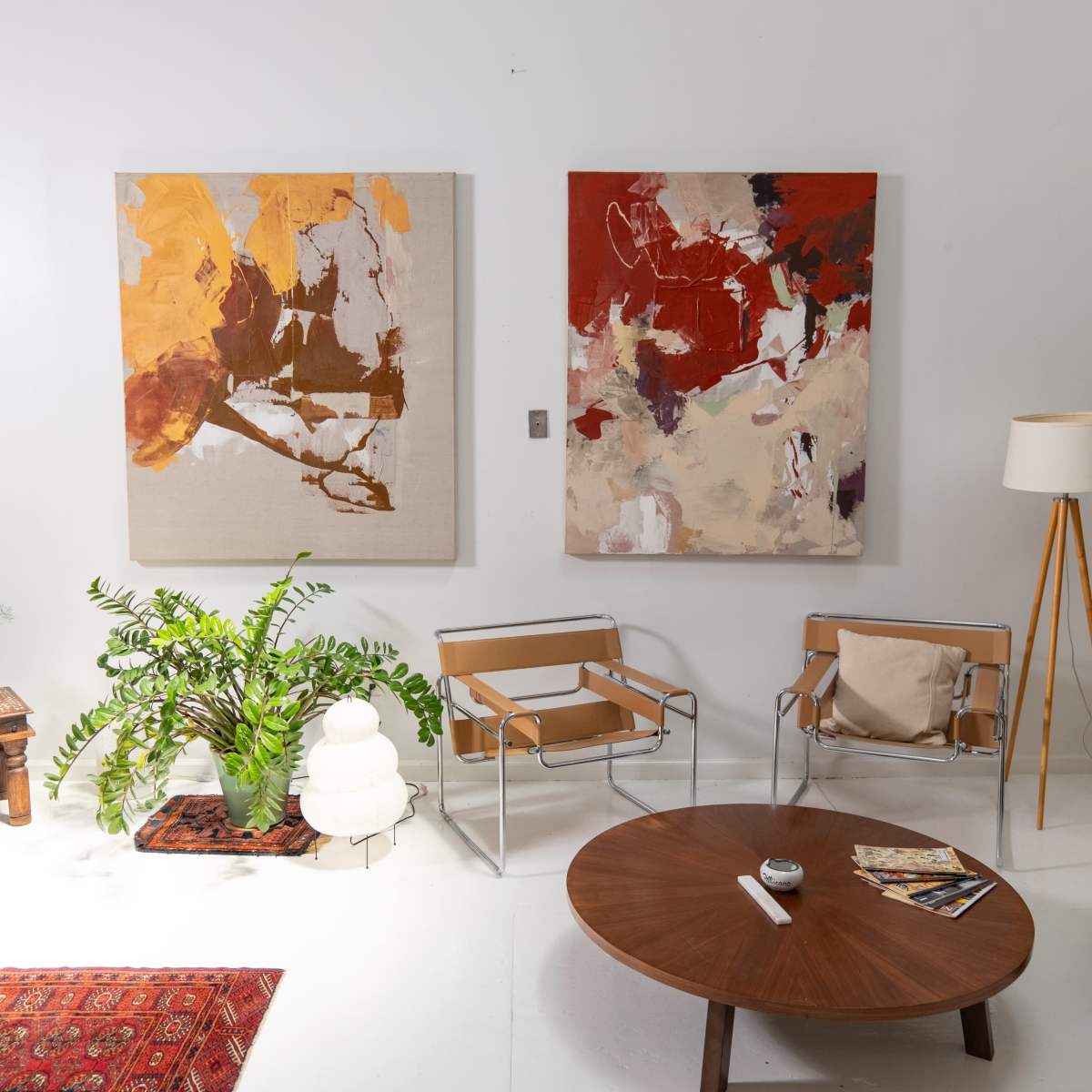A modern living room with two abstract paintings on the wall, a round wooden coffee table, minimalist chairs, and a green potted plant.