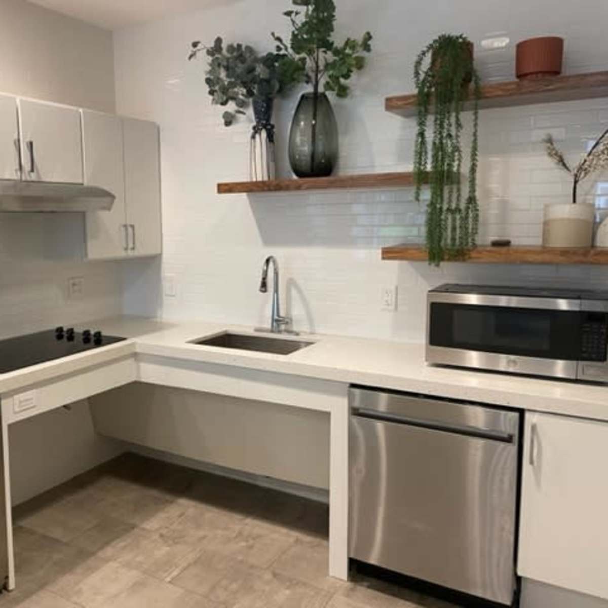 A modern kitchen with white cabinetry, a stainless steel sink and dishwasher, and wooden shelves displaying plants and decor.