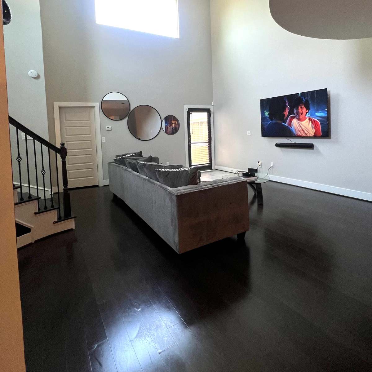 A spacious living room with a gray sofa, a large wall-mounted TV, and a staircase leading to a second floor.