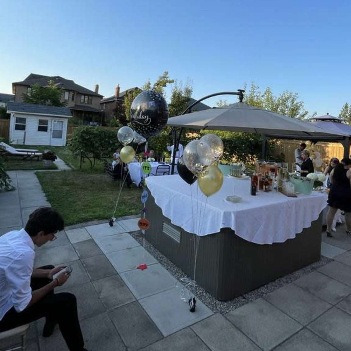 A garden setting features a table set with drinks and decorations, with guests socializing nearby under a clear blue sky.