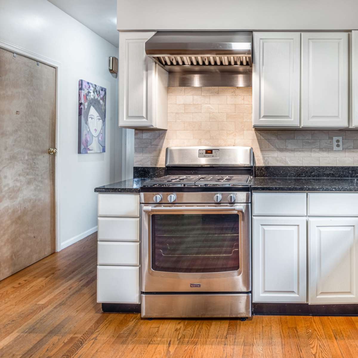 A modern kitchen features stainless steel appliances, white cabinetry, and a dark granite countertop with a wooden floor.