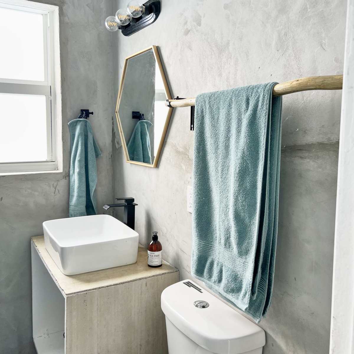 A modern bathroom featuring a concrete wall, a white square sink on a concrete vanity, a toilet, a towel rack with blue towels, and a hexagonal mirror above the sink.