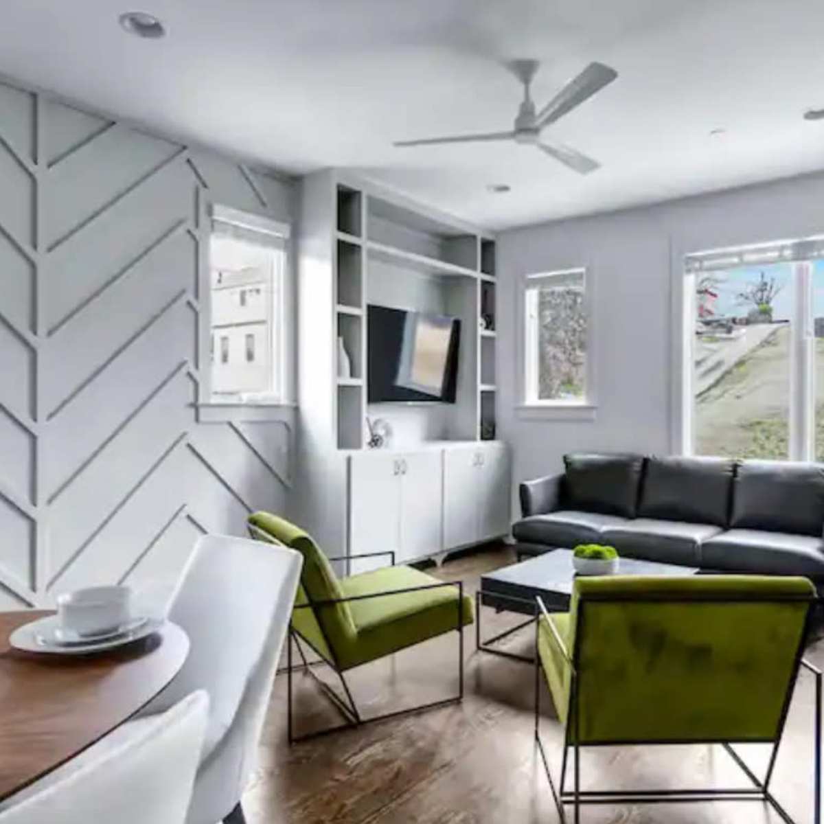 A modern living room with a black sofa, two green armchairs, a dining table set for two, and large windows that overlook a grassy area.