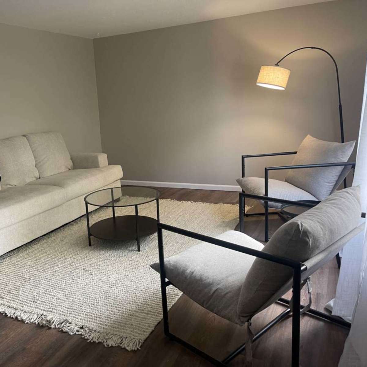A simple living room with a light sofa, two armchairs, a round glass coffee table, and a textured rug.