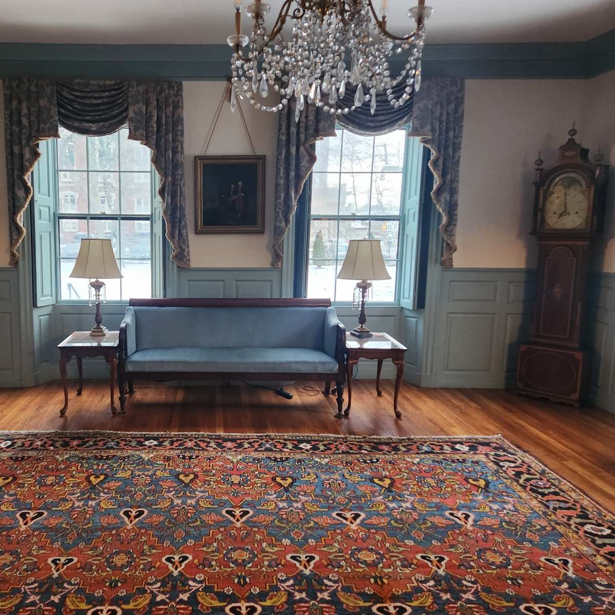 The room features a blue sofa, two lamps on side tables, a large area rug, and decorative elements including a chandelier and wall art.