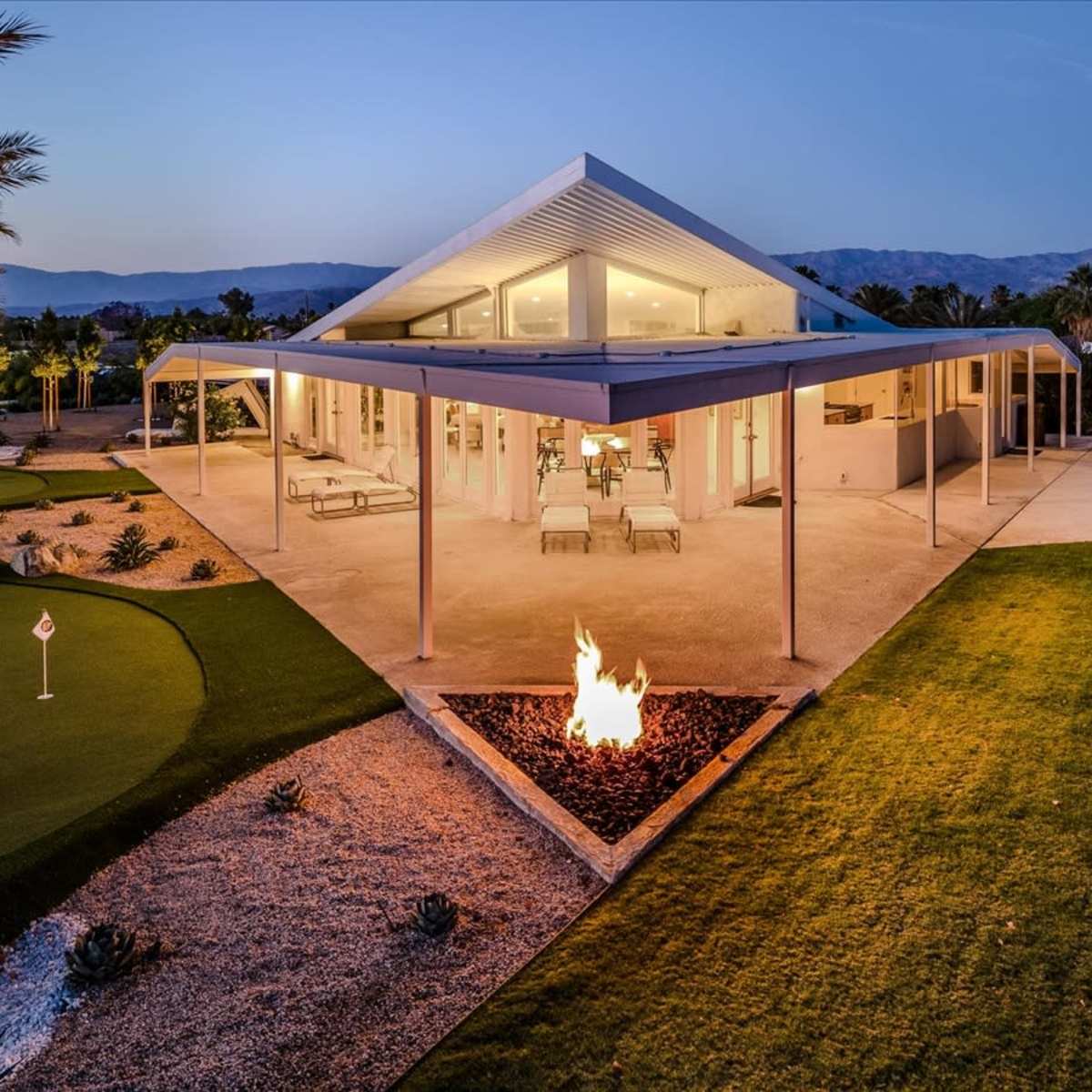 A modern house with a triangular roof and large windows is surrounded by landscaped gardens and features a fire pit in the foreground.
