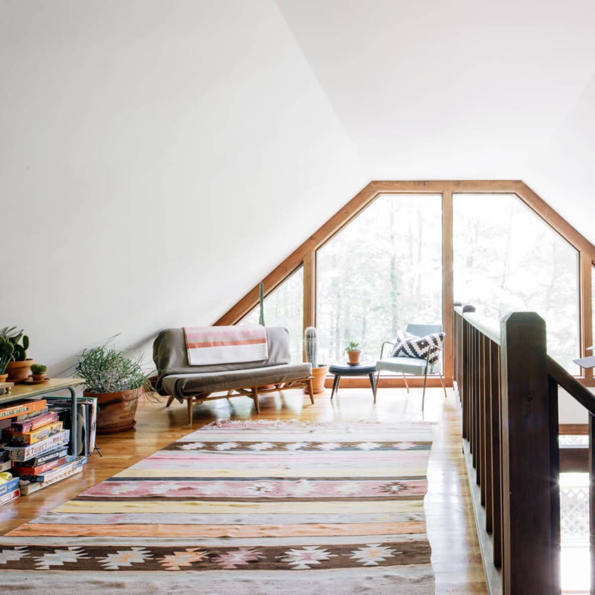 A bright, triangular attic space with large windows overlooking greenery, furnished with a couch and decorative rugs.