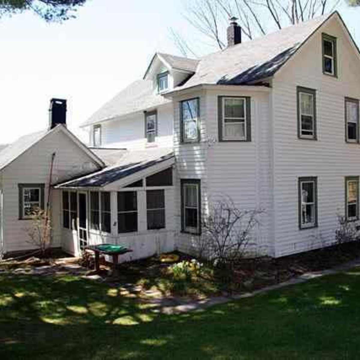 A two-story white house with a large porch and several windows stands in a grassy area, surrounded by trees and a scenic background.