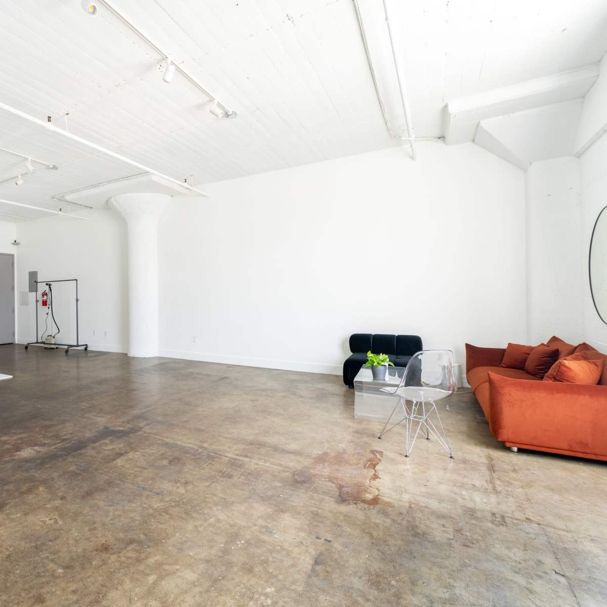 A spacious, minimalist studio with a concrete floor, a white backdrop, and a modern orange couch accompanied by a clear acrylic chair.