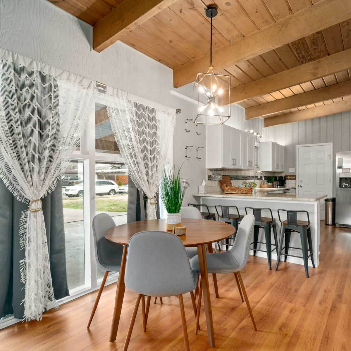 A modern kitchen and dining area featuring a round table with gray chairs, large windows, and wood-beamed ceilings.