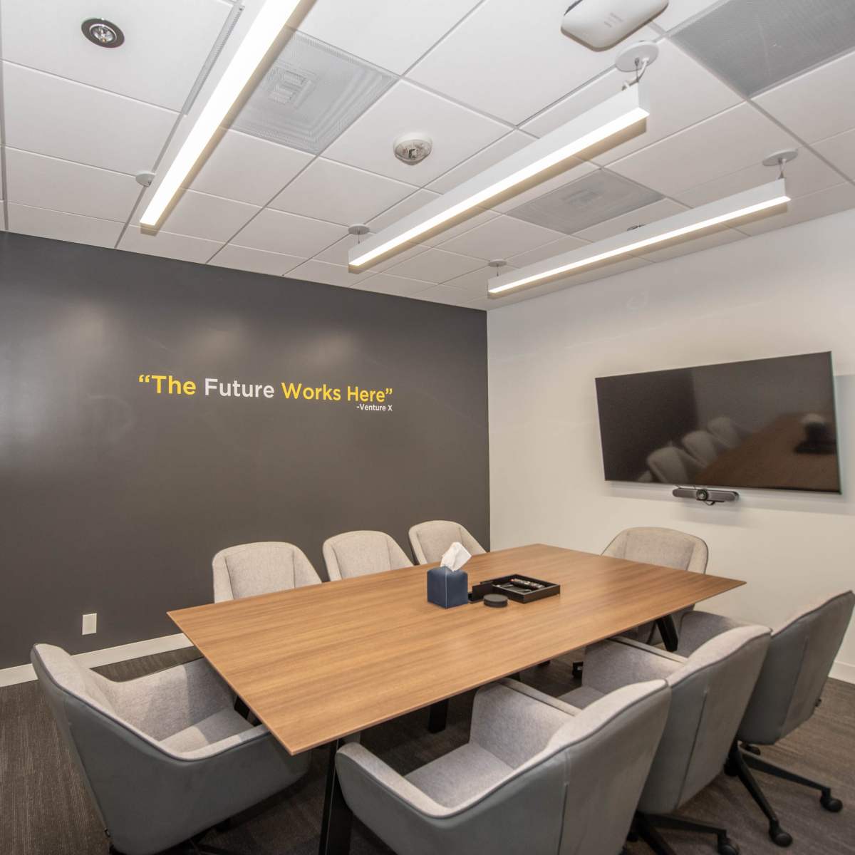 A conference room features a large wooden table surrounded by six gray chairs, with a wall displaying the phrase "The Future Works Here" and a mounted TV.