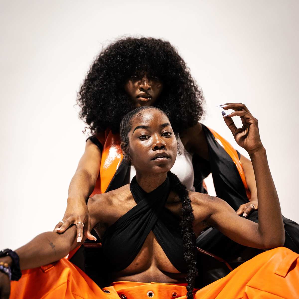 Two individuals pose together, with one sitting in front wearing a black halter top and orange pants, and the other standing behind, draped in a black outfit.