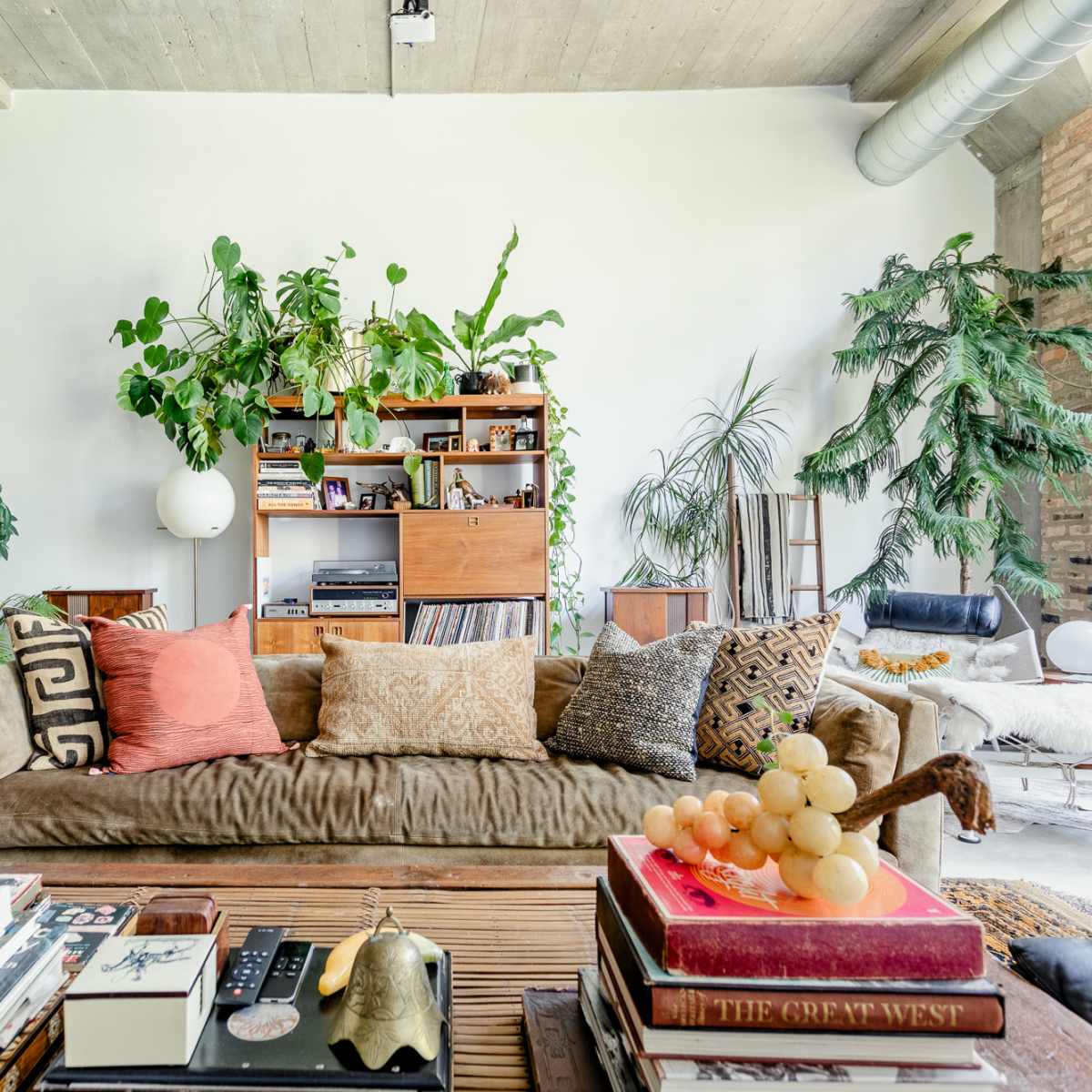 A spacious living room features a large, brown sofa adorned with various decorative pillows, surrounded by plants and a bookshelf filled with books and decor items.